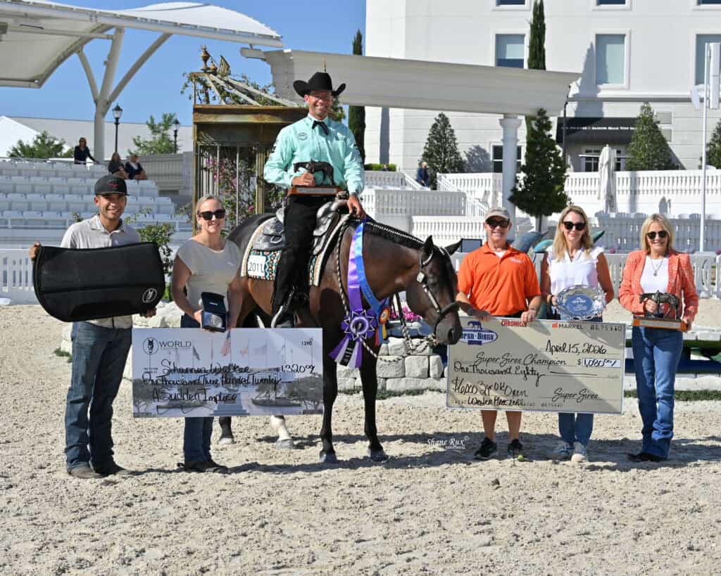 A group of six people, including a person on a horse with a ribbon, pose outdoors holding prize checks, plaques, and awards at an equestrian event.
