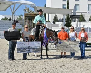 A group of six people, including a person on a horse with a ribbon, pose outdoors holding prize checks, plaques, and awards at an equestrian event.