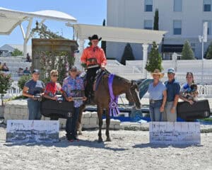 A group of people, including a person on horseback with a purple ribbon, pose outdoors with large checks and cases at an equestrian event.