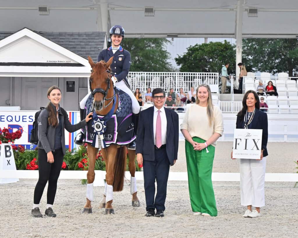 A smiling equestrian sits on a horse, holding a ribbon, flanked by four people standing on a dressage arena with event signage in the background.