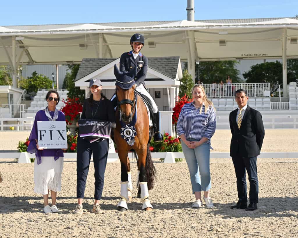 Five people and a horse pose for a photo at an outdoor equestrian event. One person is mounted on the horse, while the others stand on the ground holding signs and awards.