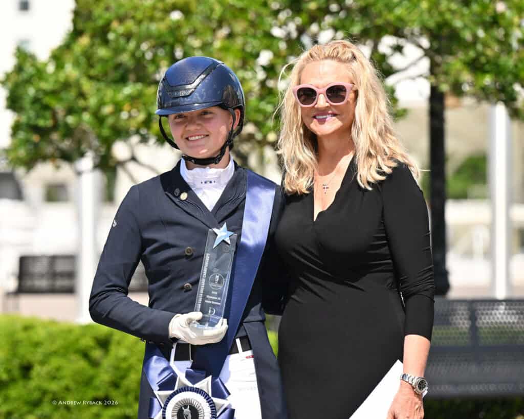 A young equestrian in riding attire and helmet holds a trophy and ribbon while standing next to a smiling woman in sunglasses and a black dress outdoors.
