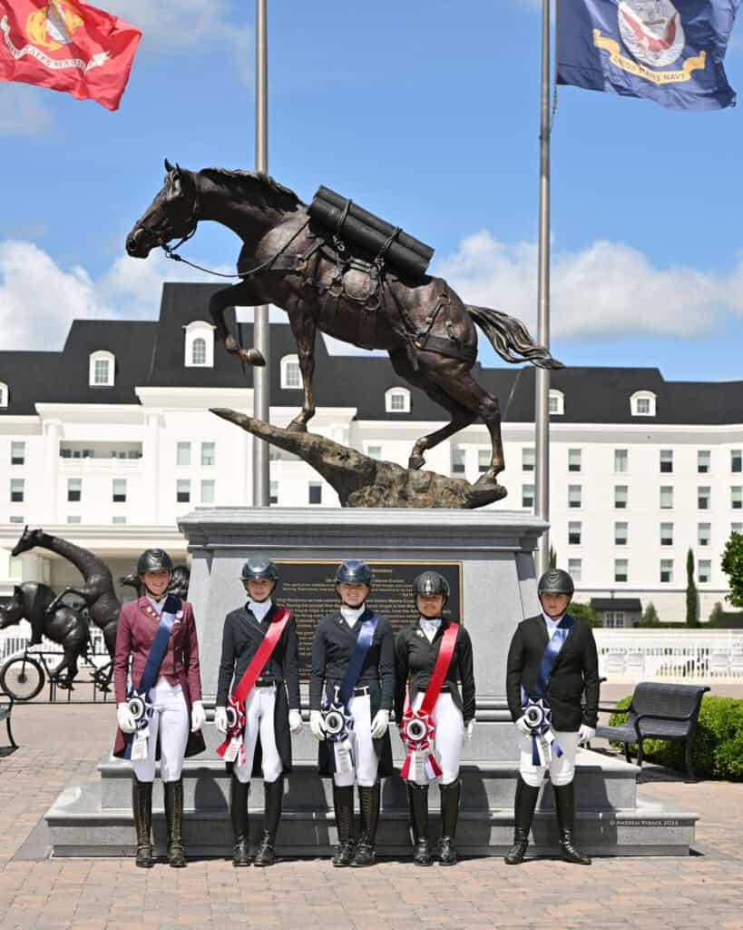 Five equestrians in riding attire stand in front of a large horse statue and plaque, holding rosettes, with flags and a building in the background.