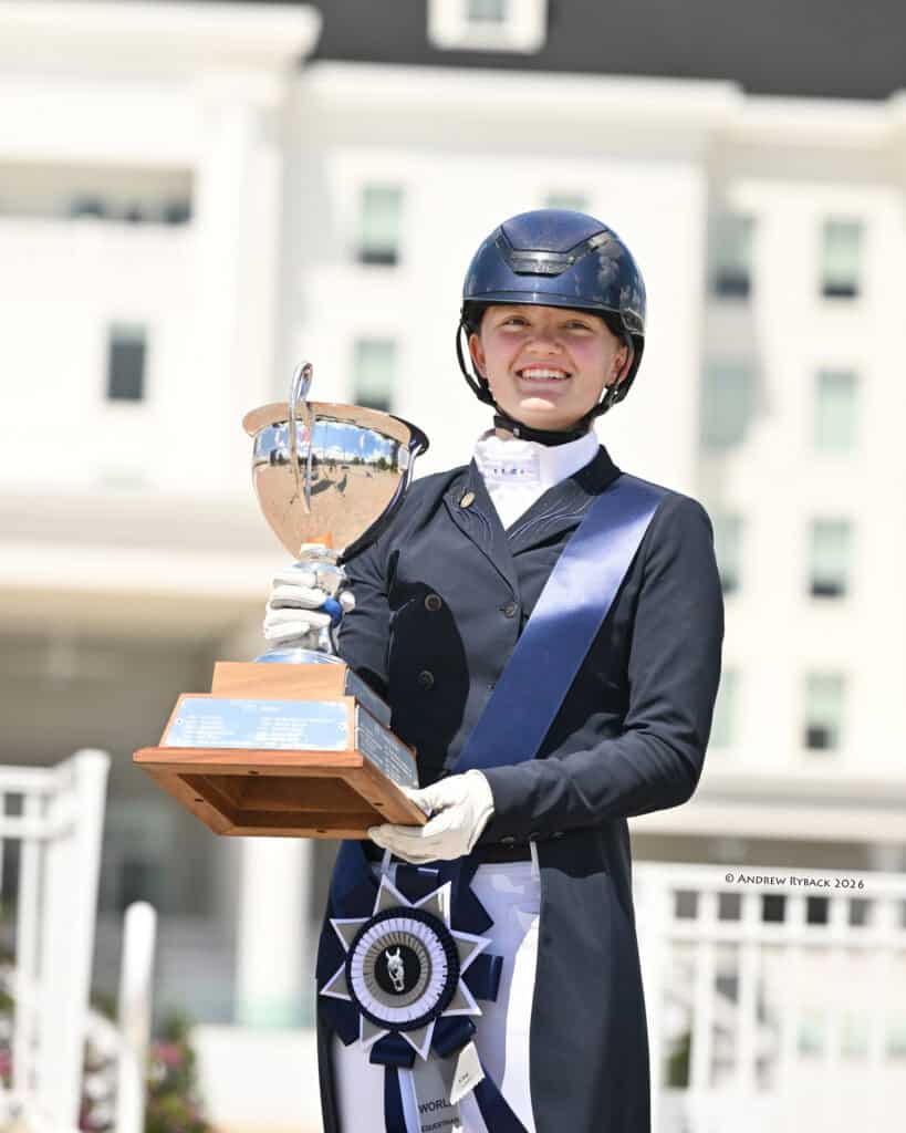 A smiling equestrian in formal riding attire and helmet holds a large trophy and a blue ribbon, standing outdoors in front of a building.