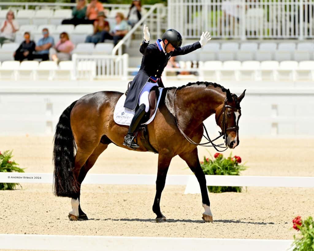 Equestrian rider in formal attire raises both arms while sitting on a brown horse during a dressage event in an arena with spectators in the background.