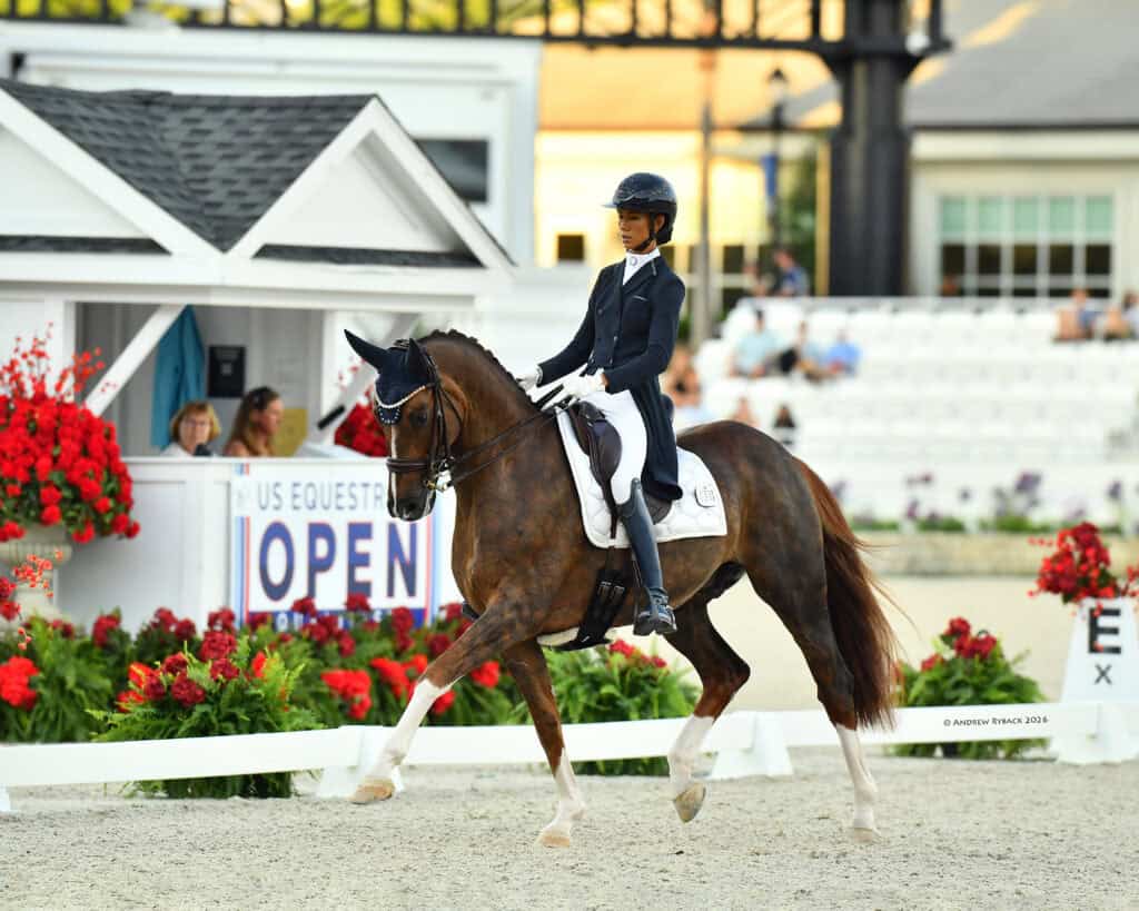 A rider in formal equestrian attire guides a horse during a dressage competition, with an "OPEN" US Equestrian sign and red flowers in the background.