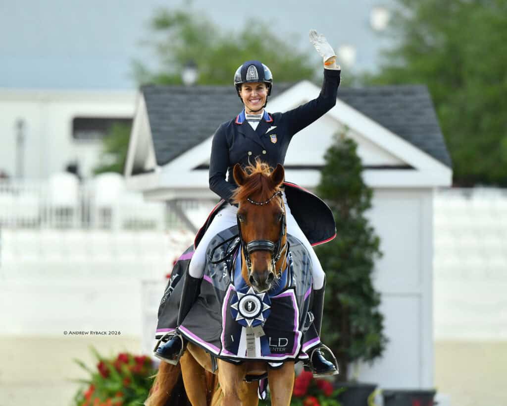 Equestrian rider in formal attire waves while sitting on a horse draped in a winner’s blanket at an outdoor competition.