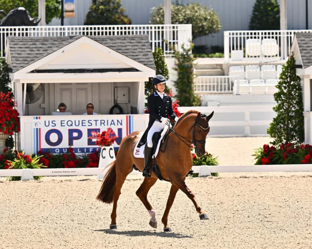 A rider in formal attire guides a horse in a dressage arena at the US Equestrian Open, with a white judge’s booth and event signage in the background.