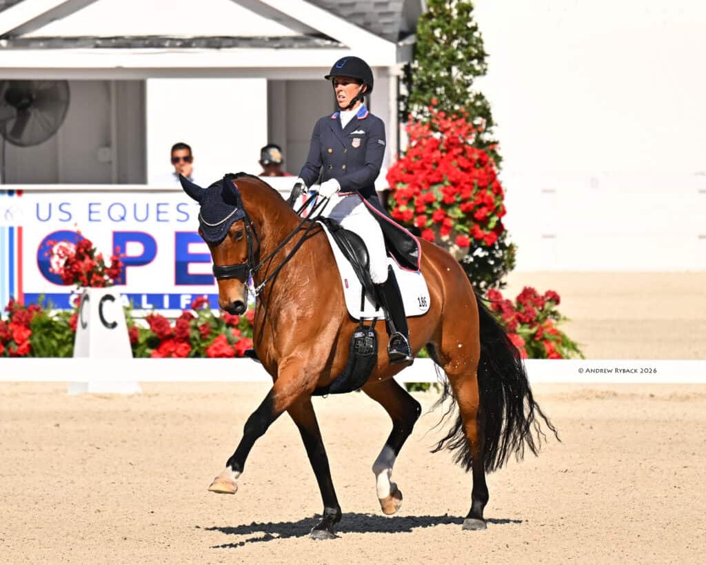 A rider in formal equestrian attire guides a brown horse in a dressage arena, with red flowers and a US Equestrian sign visible in the background.