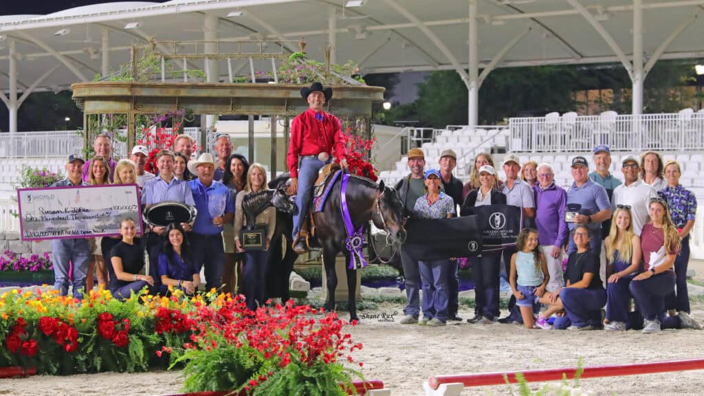 A group of people pose around a person on horseback at an arena, holding awards, plaques, and an oversized check, with flowers and decorations in the foreground.