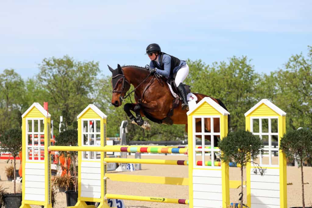 A rider in equestrian gear jumps a brown horse over a yellow and white obstacle during a show jumping competition outdoors.