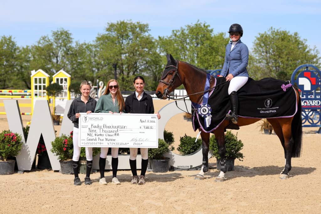Four people stand holding a large check next to a person on a horse wearing a winner’s sash and ribbon at an outdoor equestrian event.