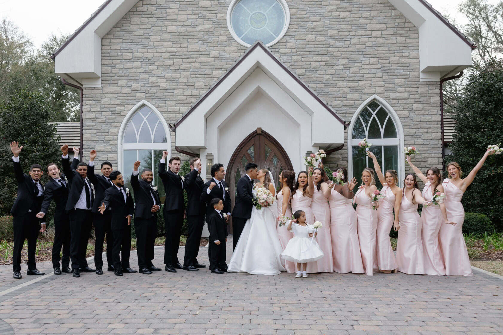 A bride and groom stand with their wedding party in front of a stone church; bridesmaids wear pale pink dresses and groomsmen wear black suits.
