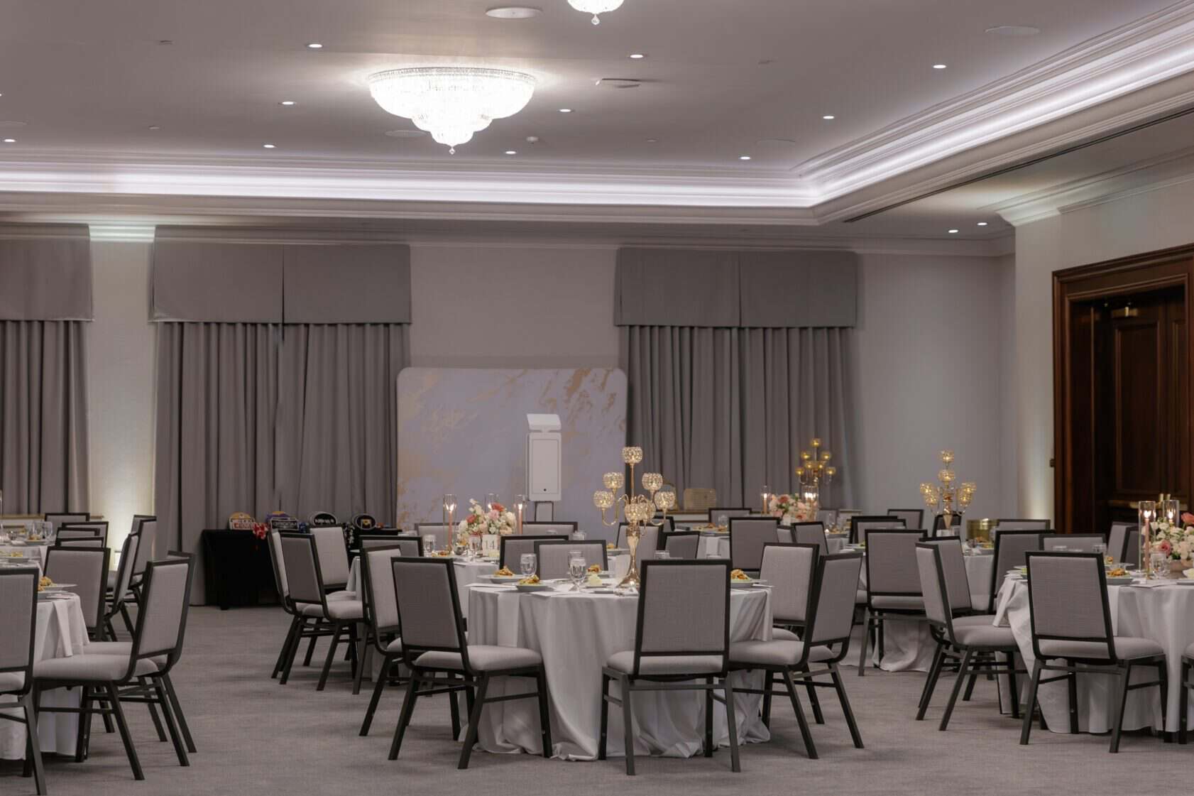 A banquet hall with round tables and gray chairs, set for an event with floral centerpieces and neutral decor, under a large chandelier.