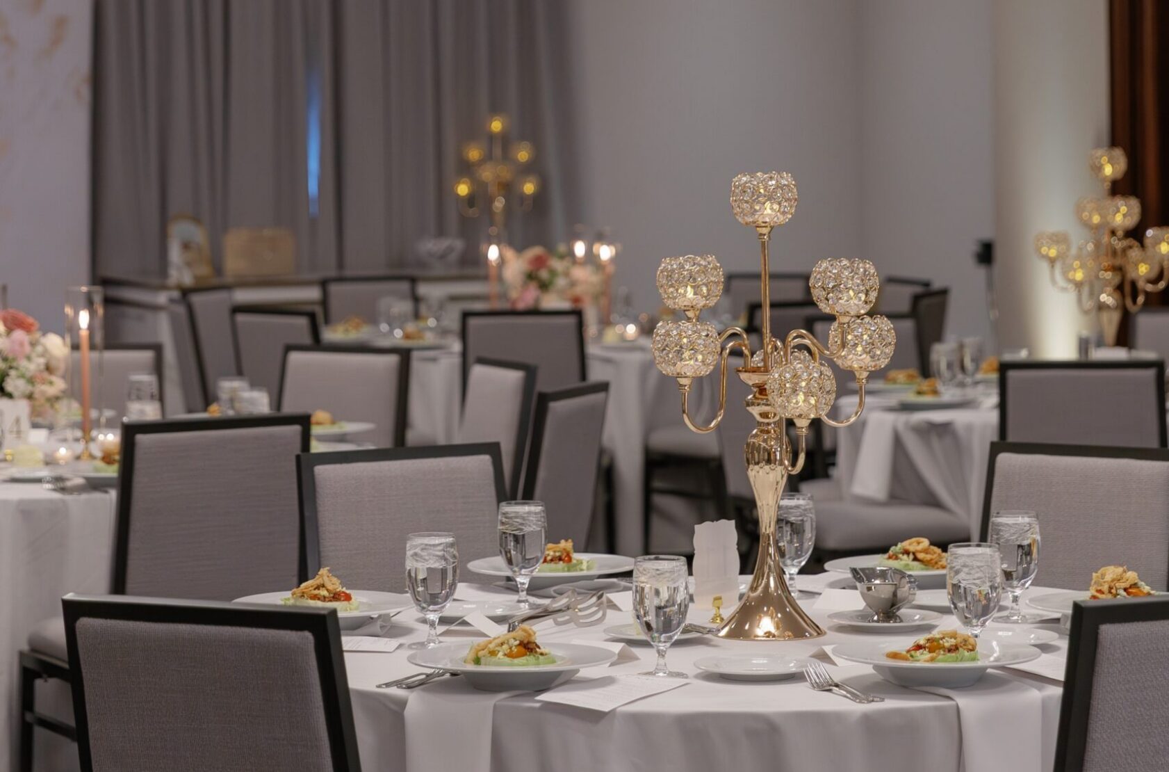 A banquet hall set with round tables, white tablecloths, grey padded chairs, gold candelabras, and plates of food, ready for a formal event.