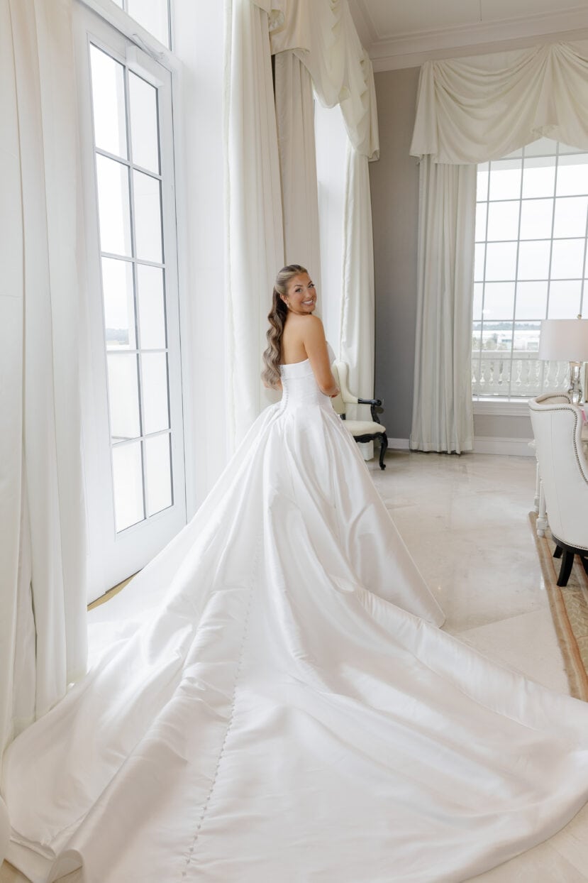 A woman in a strapless white wedding gown with a long train stands indoors near large windows, looking over her shoulder and smiling.