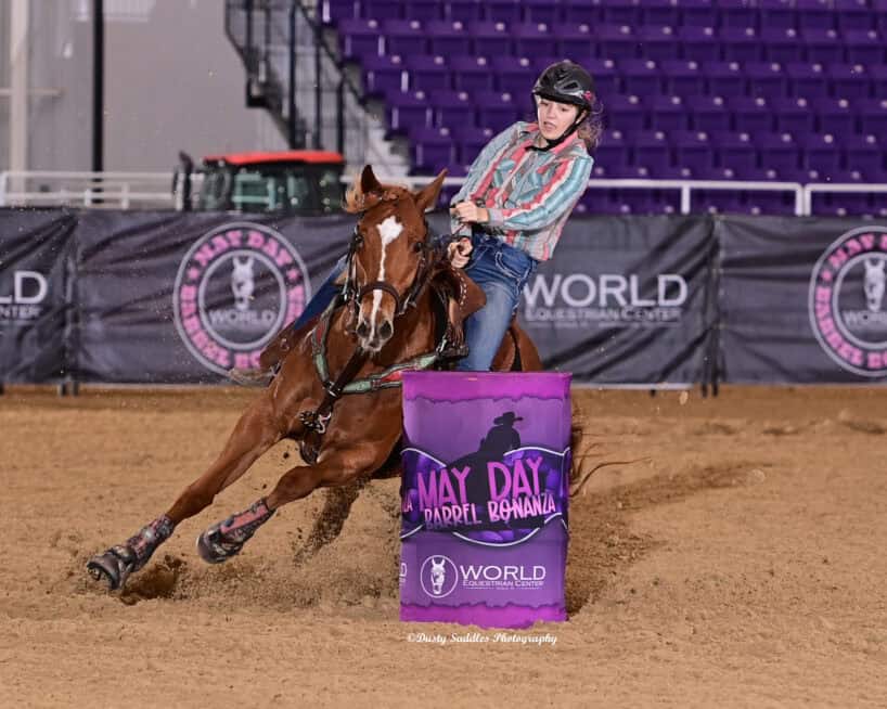 A rider wearing a helmet and plaid shirt guides a horse sharply around a purple barrel in an indoor arena during a barrel racing event.