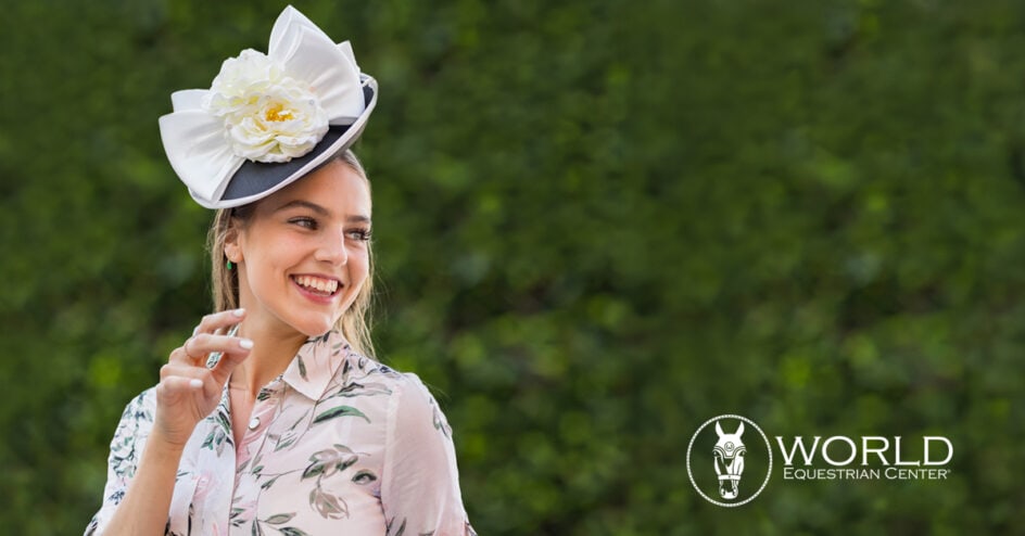A woman wearing a floral dress and an elaborate white hat smiles outdoors; the World Equestrian Center logo appears in the bottom right corner.