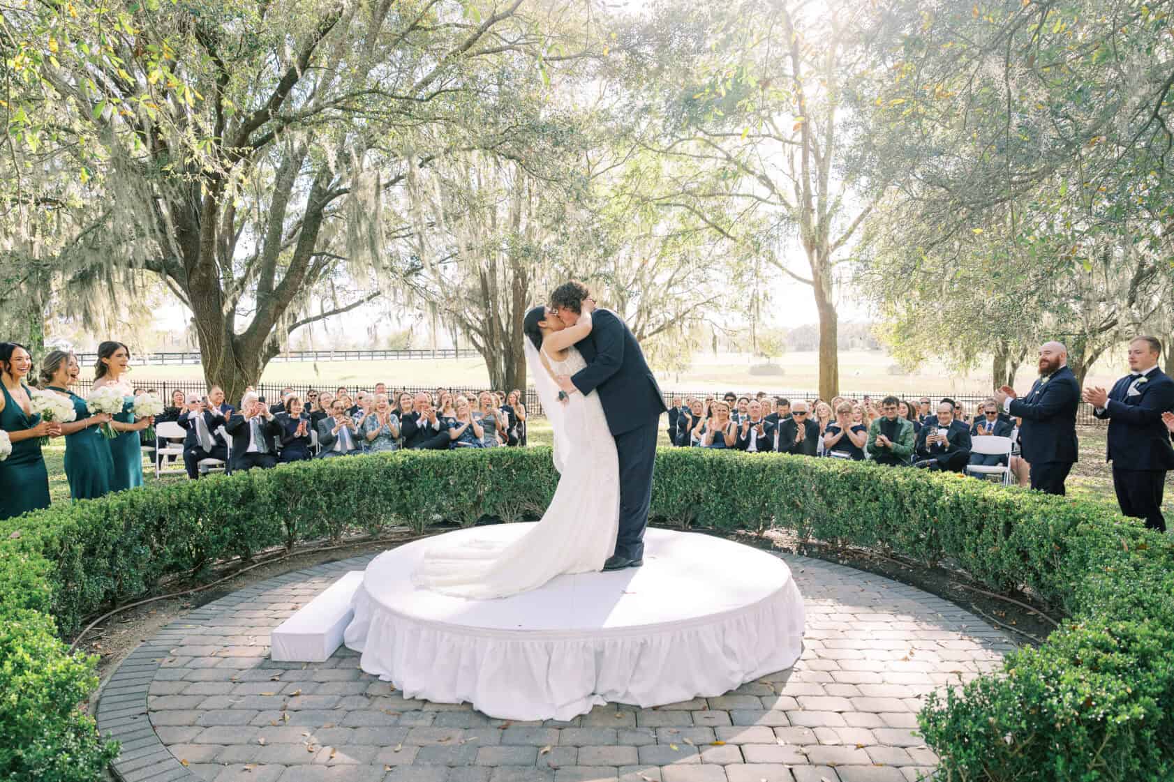 A bride and groom kiss on a round platform at an outdoor wedding ceremony, surrounded by guests seated in rows and sunlight filtering through trees.