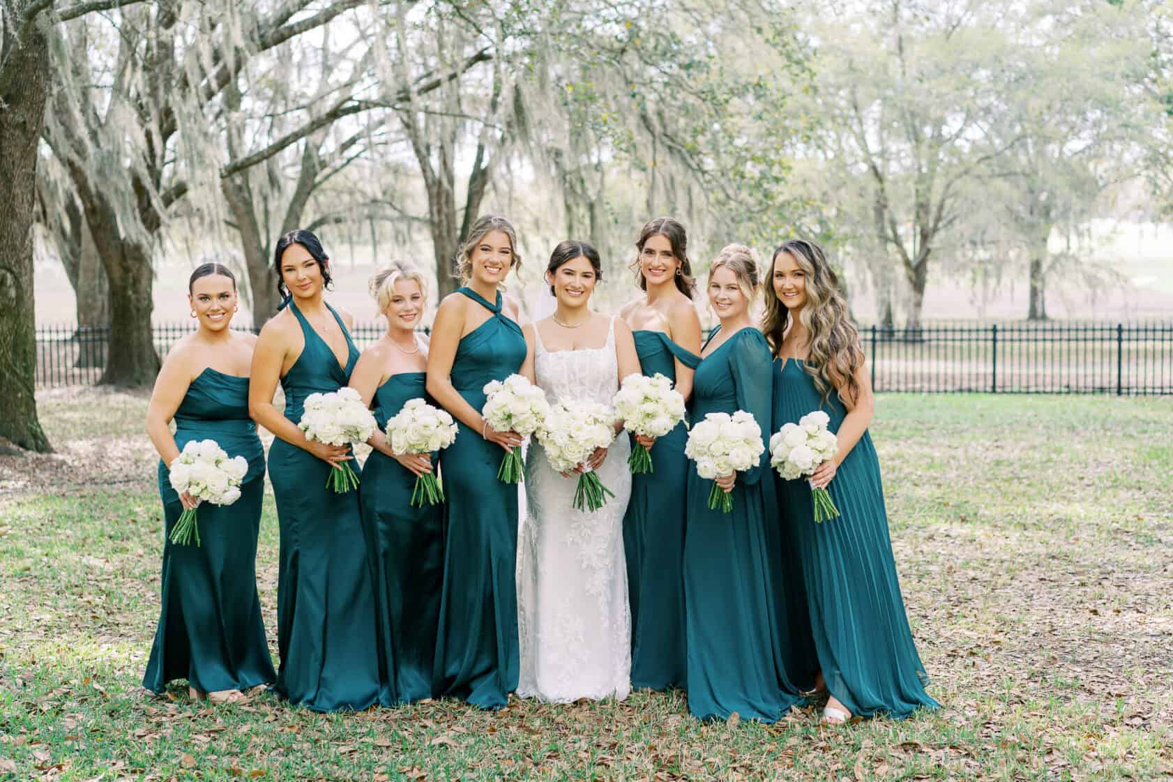 Eight bridesmaids in teal dresses and one bride in white stand outdoors in a row, all holding white flower bouquets, with trees and a black fence in the background.