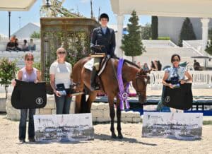 Four women at an equestrian event; one on horseback wearing riding attire with a ribbon, and three holding oversized checks and awards in an outdoor arena setting.