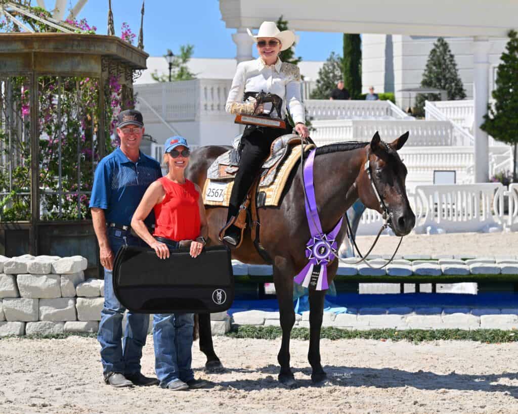 A woman in show attire sits on a horse with a large purple ribbon. Two people stand beside her holding an award plaque. All are outdoors in a sunny equestrian arena.