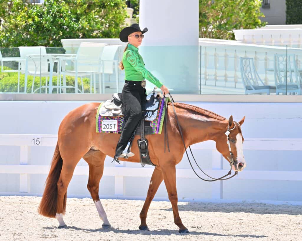 A rider in a green shirt and black hat is riding a brown horse with a white blaze on its nose in an outdoor arena.