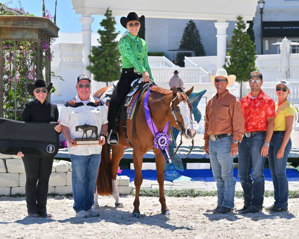 A group of six people poses outdoors; one person sits on a horse with a large purple ribbon, while others stand holding awards and wearing cowboy hats.