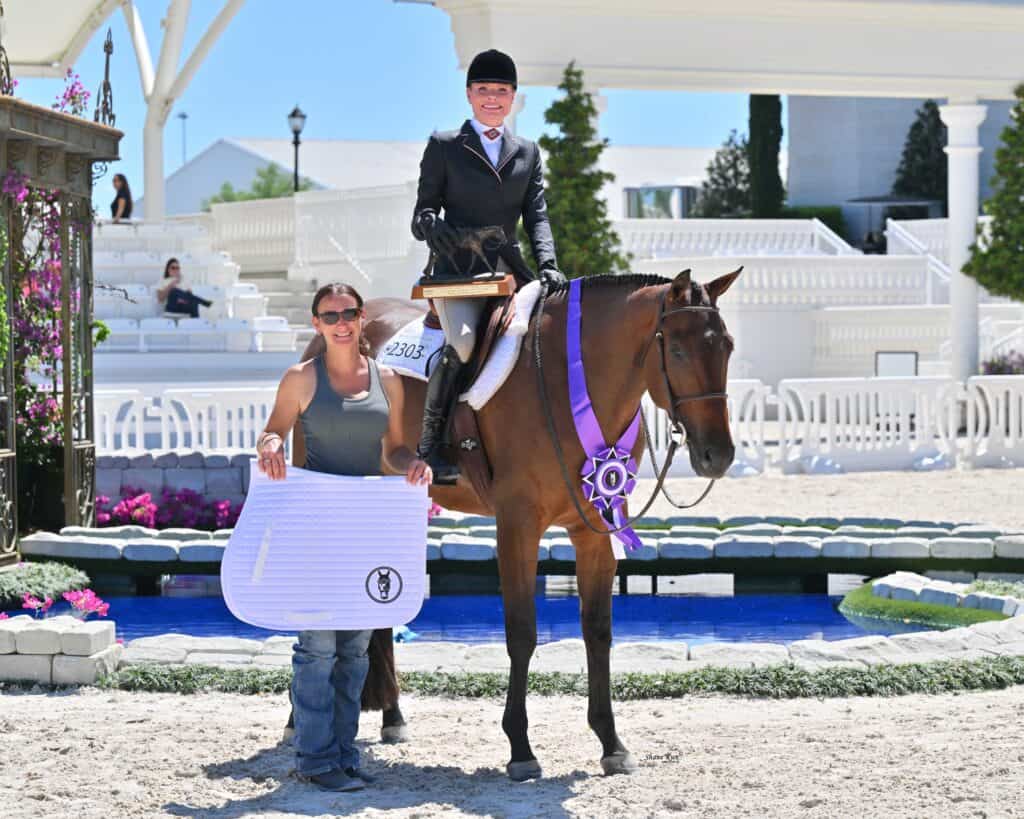 Two women pose with a horse in an outdoor arena; one is mounted and holding a trophy, the other stands beside them holding a saddle pad, with a large purple ribbon on the horse's shoulder.