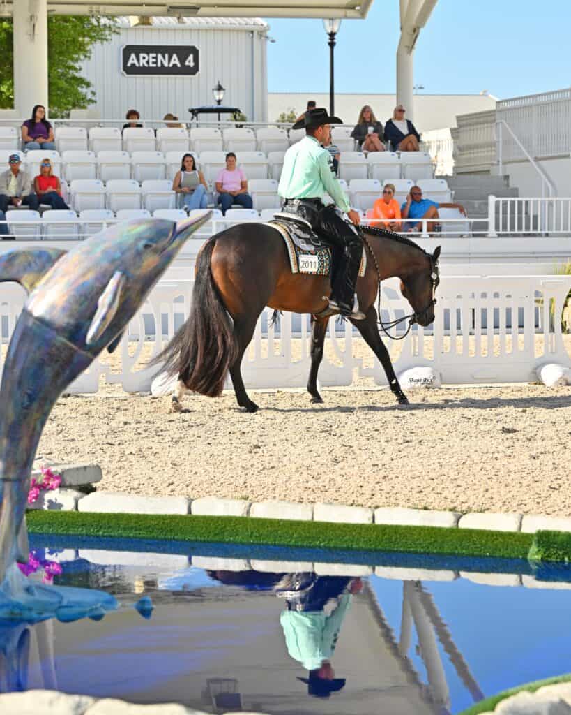 A person in western attire rides a horse in a sandy arena with spectators watching; a dolphin sculpture and reflecting pool are in the foreground.