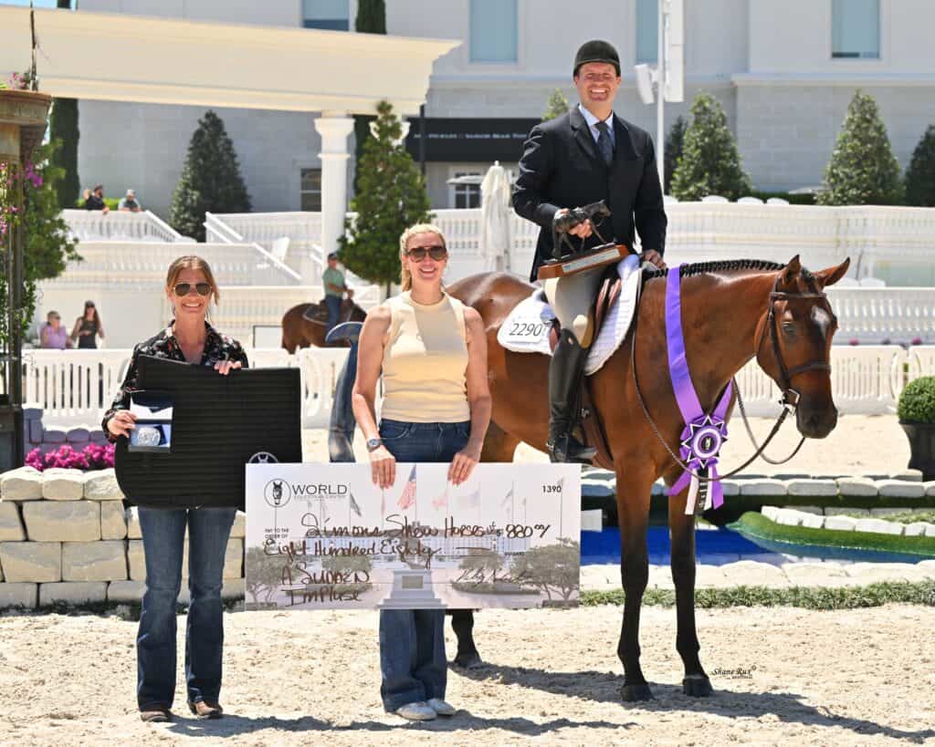 Three people and a horse pose outdoors; one person on horseback holds purple ribbons, while the others hold a large check and a case, in front of a building with white fencing and landscaping.