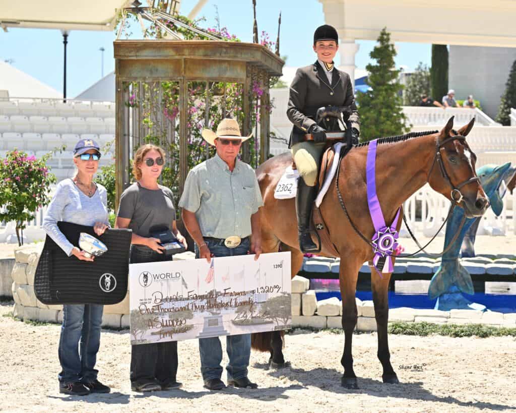 Four people and a horse pose outdoors after an equestrian event; one person holds a large check, another holds a black pad, and the horse wears a winner's ribbon.
