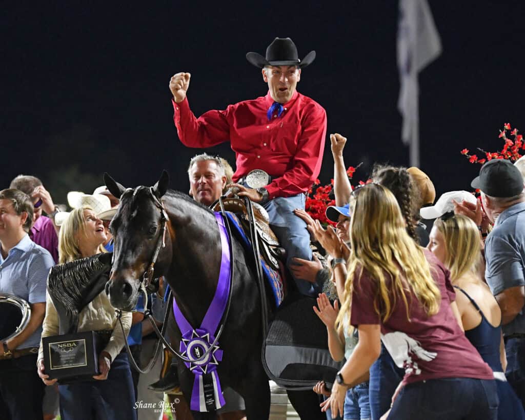 A man in a red shirt and black hat celebrates on horseback with a large purple ribbon, surrounded by cheering people at an outdoor event.