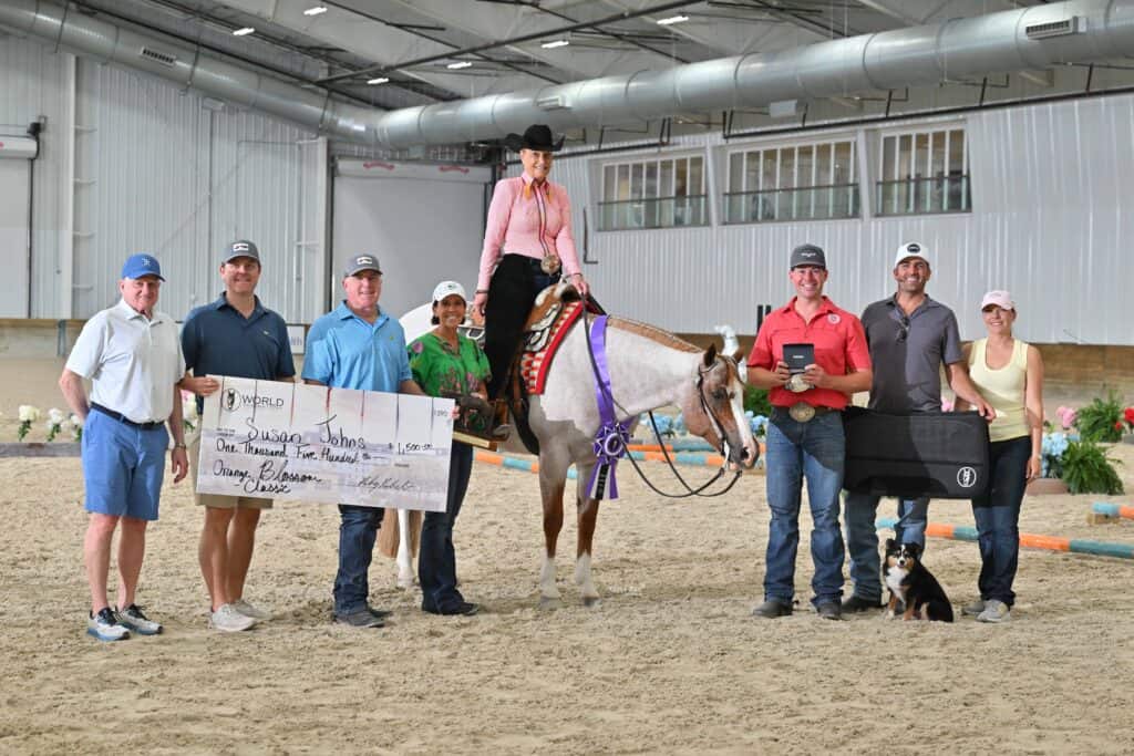 A group of people stands in an indoor arena with a horse and rider; one person holds a large check and another holds a trophy, with a dog sitting in front of the group.