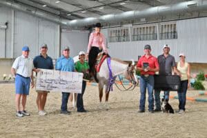A group of people stands in an indoor arena with a horse and rider; one person holds a large check and another holds a trophy, with a dog sitting in front of the group.