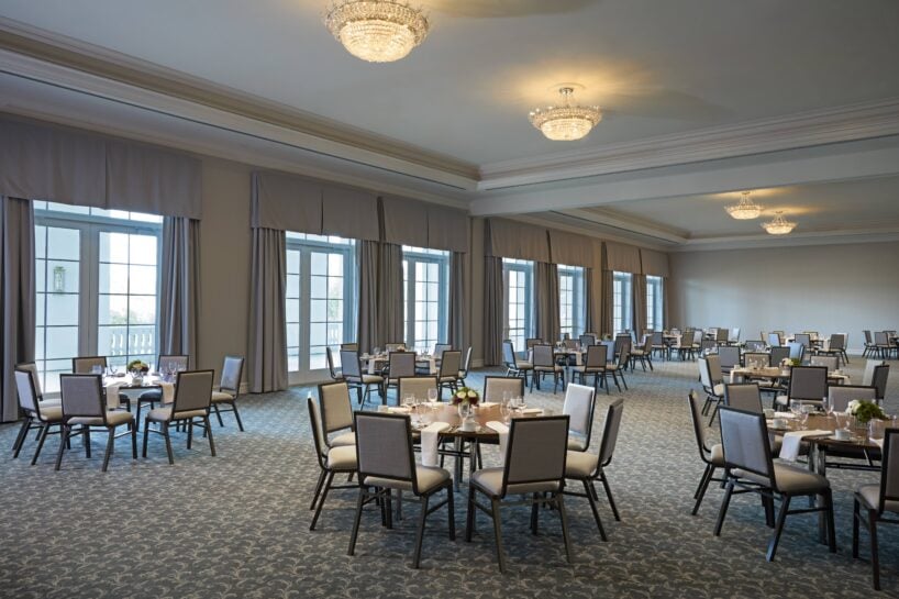 Spacious banquet hall with round tables and chairs arranged throughout, set with plates and flowers, under large chandeliers and surrounded by tall windows with curtains.