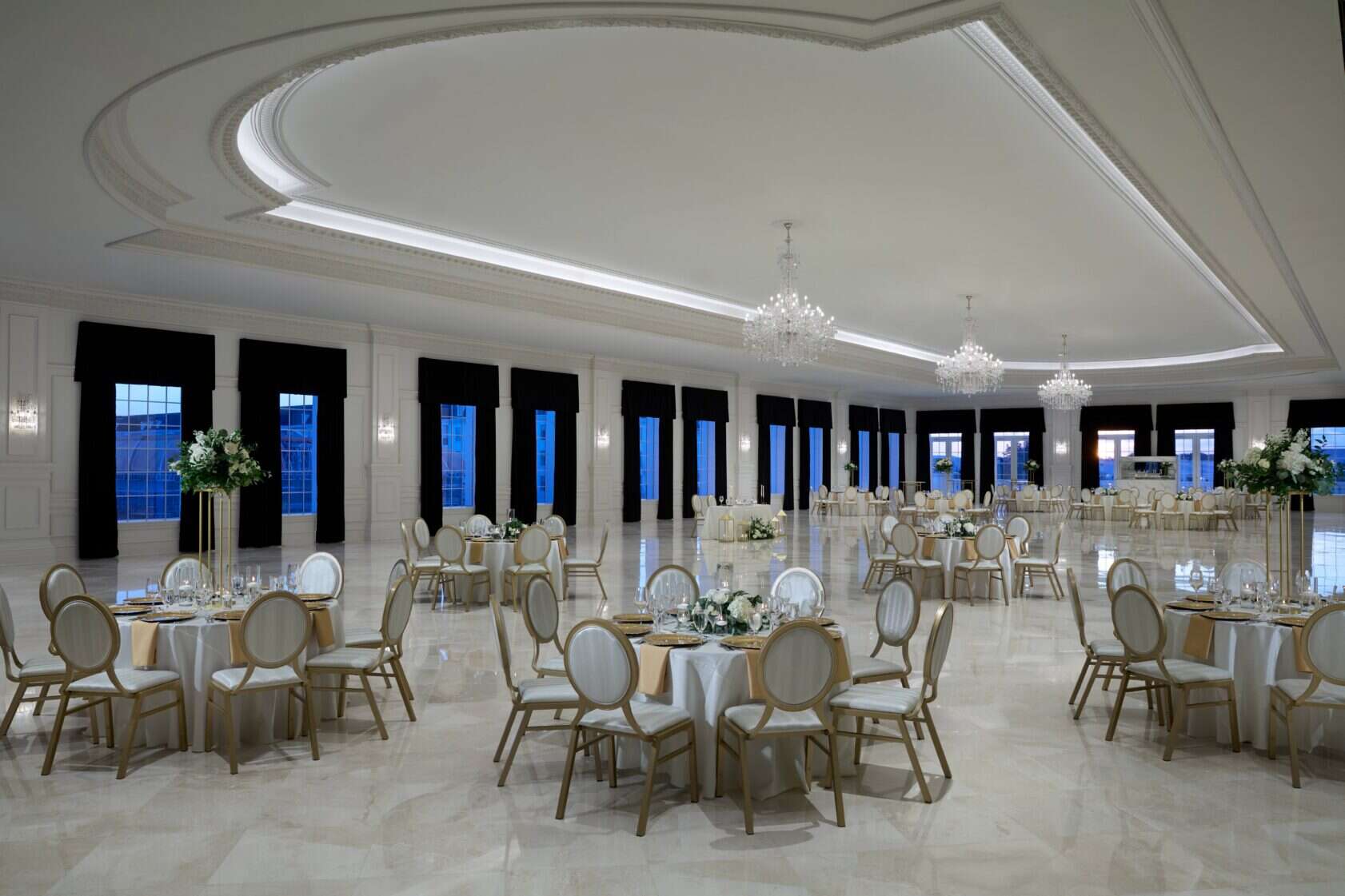 Elegant banquet hall with round tables set for an event, featuring chandeliers, large windows with black curtains, and polished marble floors.