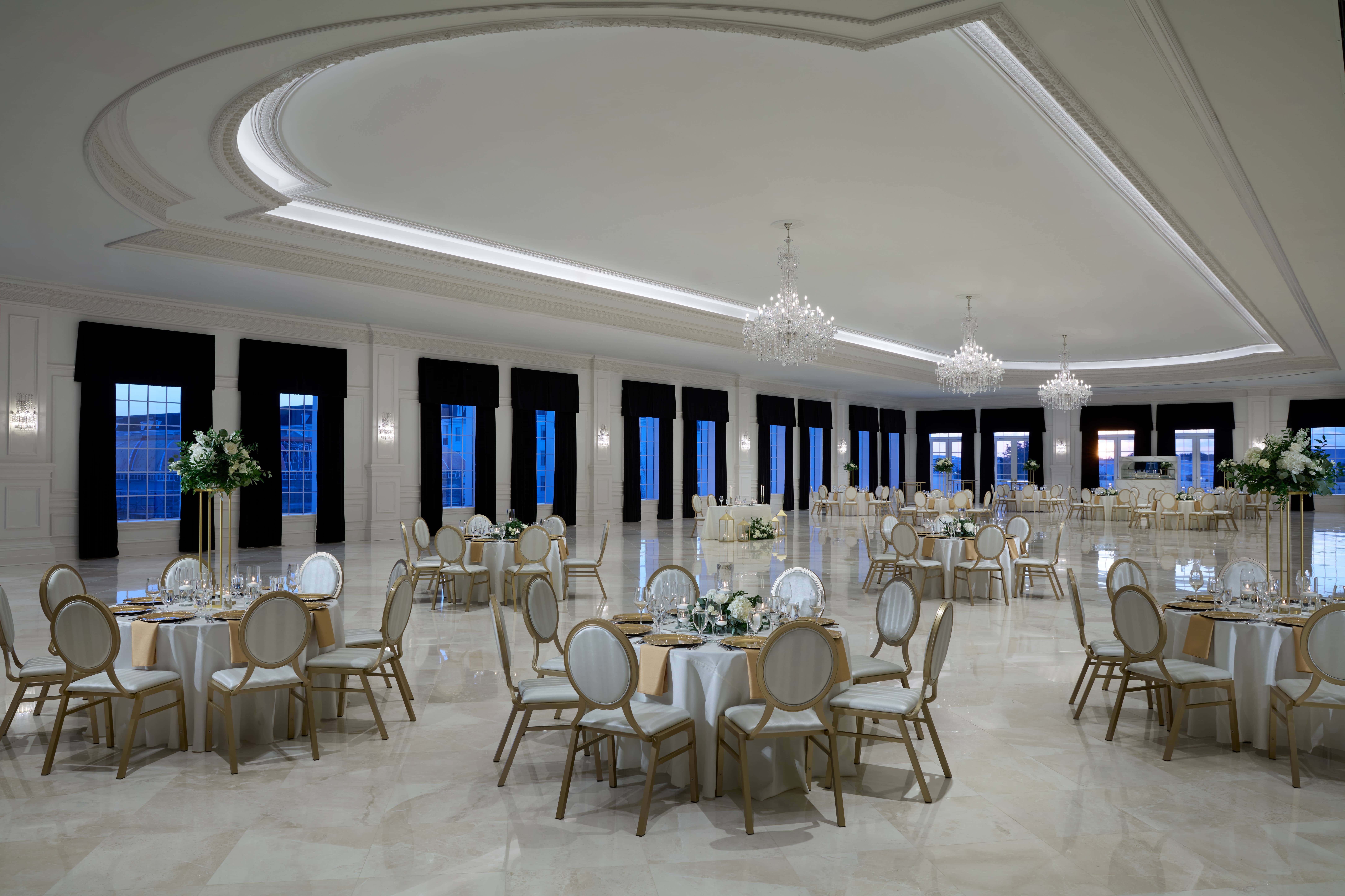 Spacious, elegant banquet hall with round tables set for an event, white and gold chairs, chandeliers, floral centerpieces, and large windows with dark curtains.