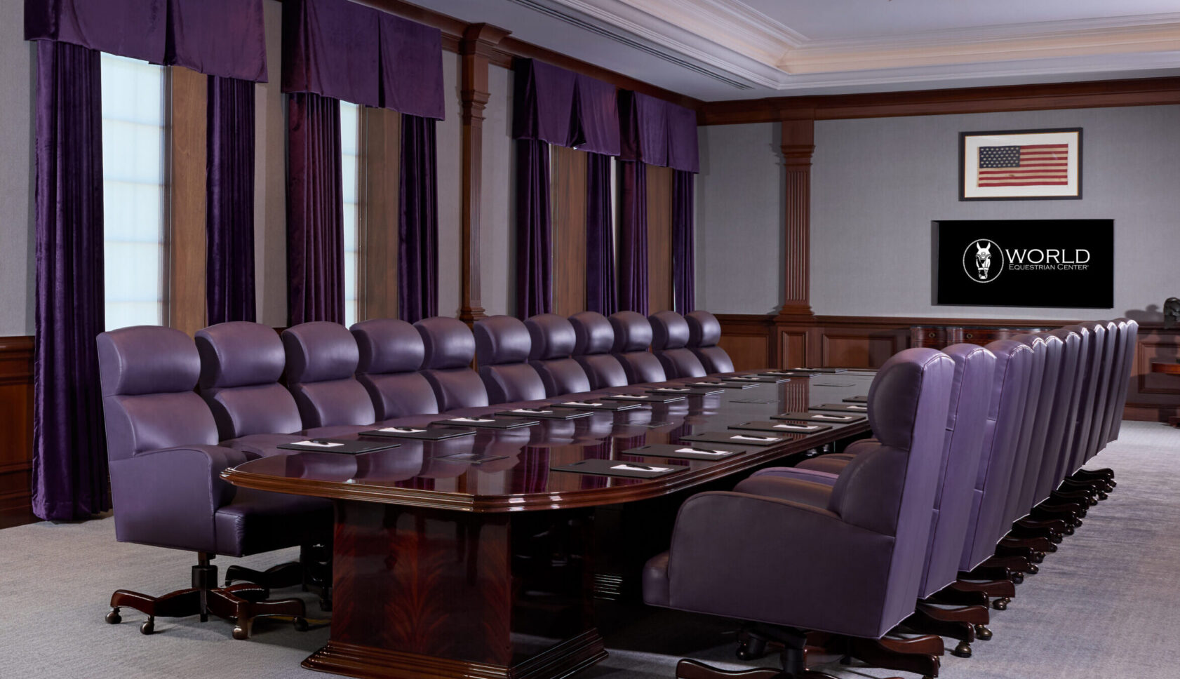 Large formal conference room with a long polished table, purple upholstered chairs, chandeliers, American flag on the wall, and a TV displaying the word "WORLD.