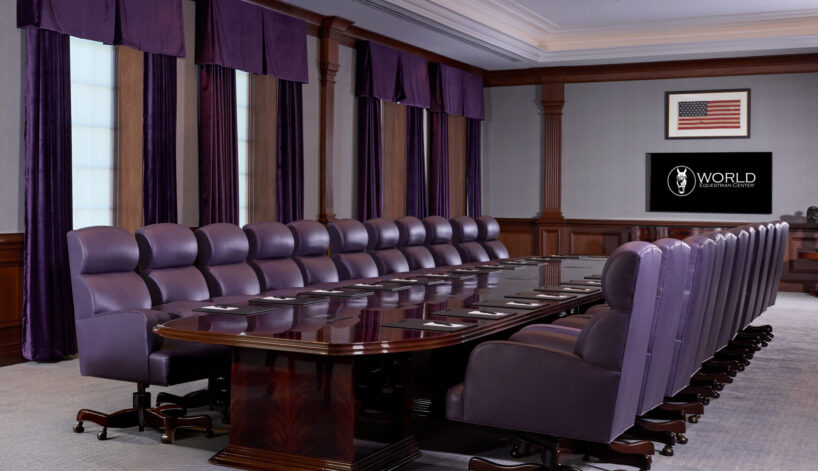 Large formal conference room with a long polished table, purple upholstered chairs, chandeliers, American flag on the wall, and a TV displaying the word "WORLD.