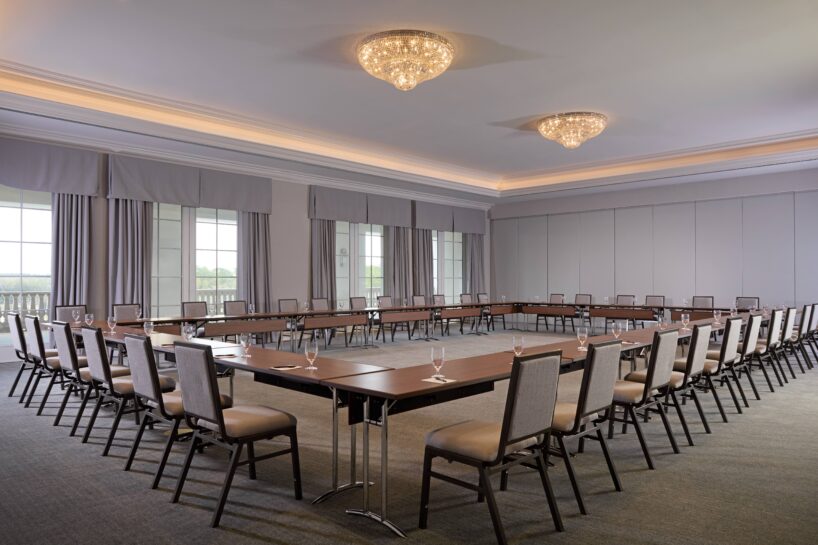 A large conference room with U-shaped tables, neatly arranged chairs, glasses of water, carpeted floors, large windows with gray curtains, and chandeliers on the ceiling.