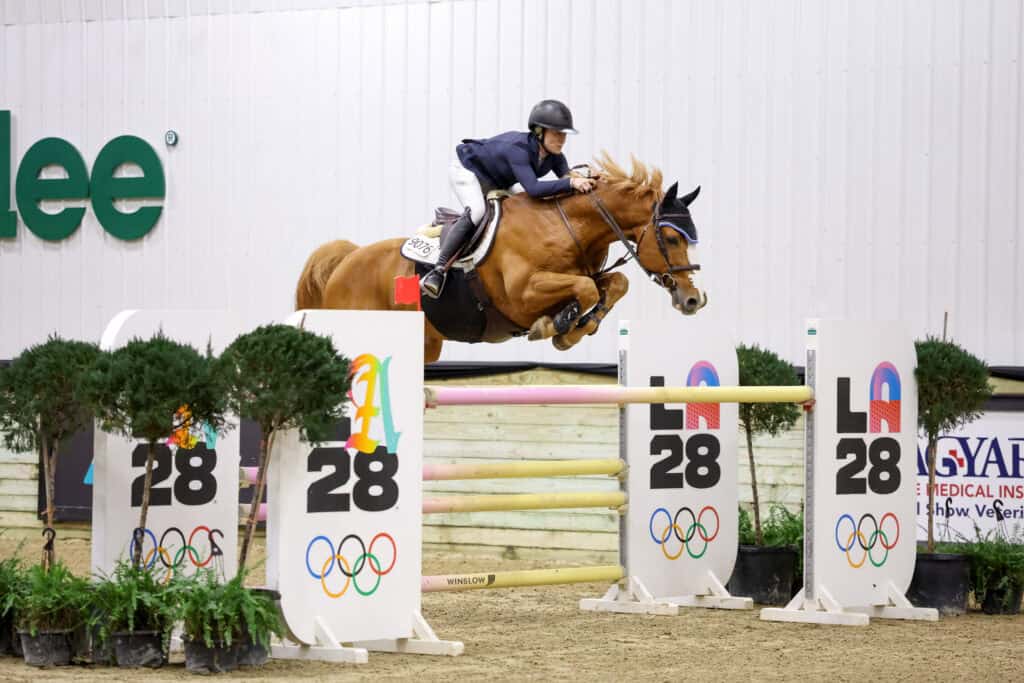 A rider on a horse jumps over an obstacle with "LA 28" and Olympic symbols in an indoor equestrian arena.