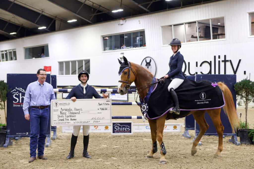 Three people pose indoors at an equestrian event; one holds a large check, and one is on a horse dressed in competition attire.