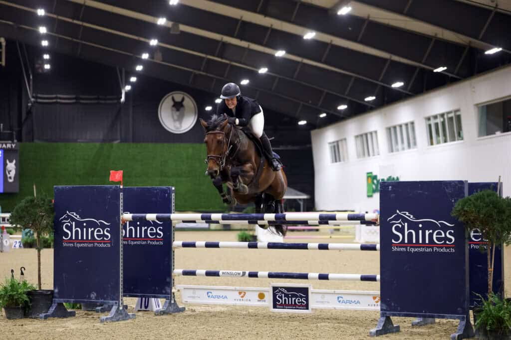A rider in equestrian gear jumps a horse over an obstacle during an indoor show jumping competition.
