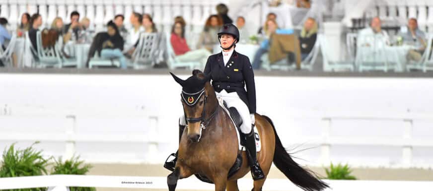 A rider in formal attire participates in an equestrian event, riding a horse in an arena with spectators seated in the background.