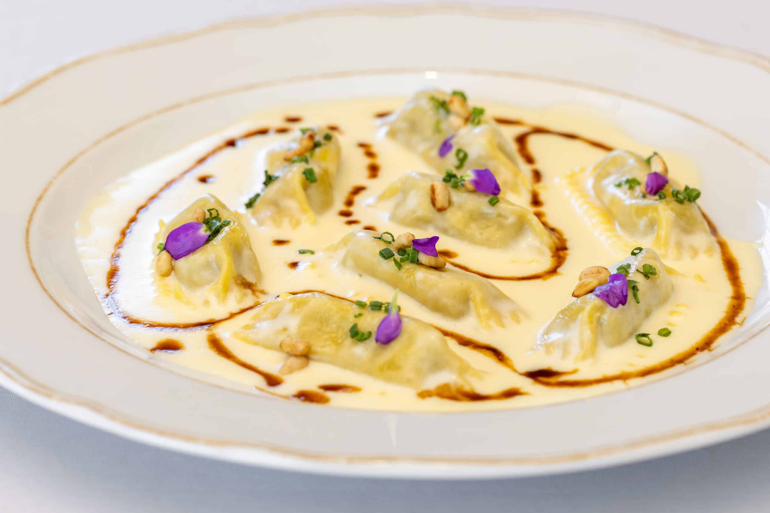 Seven dumplings in a creamy white sauce, garnished with purple edible flowers, pine nuts, chopped herbs, and a drizzle of brown sauce on a white plate.