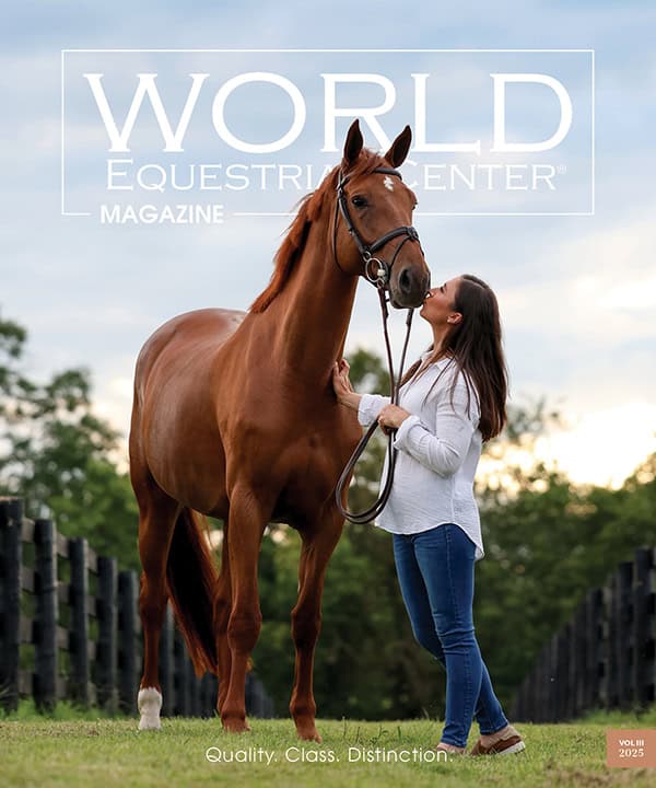 A woman in a white shirt stands beside a chestnut horse, holding its lead, on a grassy path between wooden fences with trees and a cloudy sky in the background.