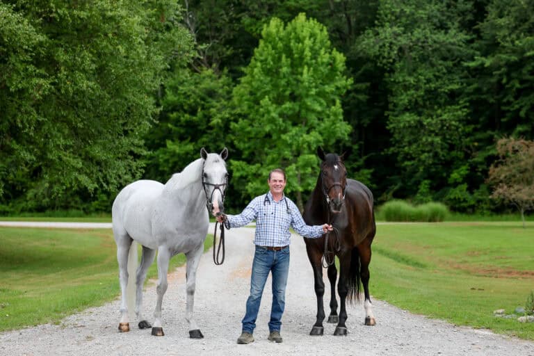 A man stands on a gravel path holding the reins of a white horse and a brown horse, with green trees and grass in the background.
