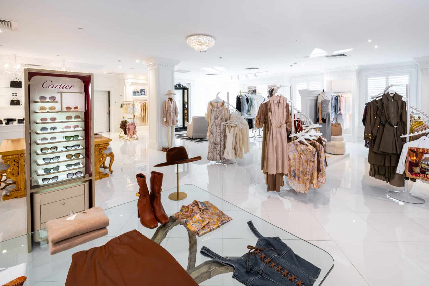 A bright, modern boutique displays women’s clothing, accessories, boots, hats, and a Cartier sunglasses case on glossy white floors.