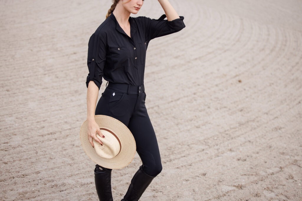 A person in a black shirt and pants stands on sandy ground, holding a wide-brimmed hat and wearing tall black boots. Their face is partially obscured by their hand.